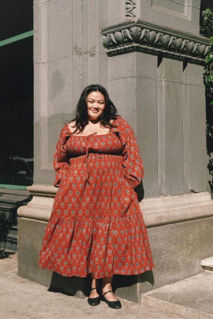 Une mannequin porte une robe rouge à motifs floraux. En fond, un bâtiment dans une ville américaine. La robe est de la marque Christy Dawn et est conçue en coton régénératif.