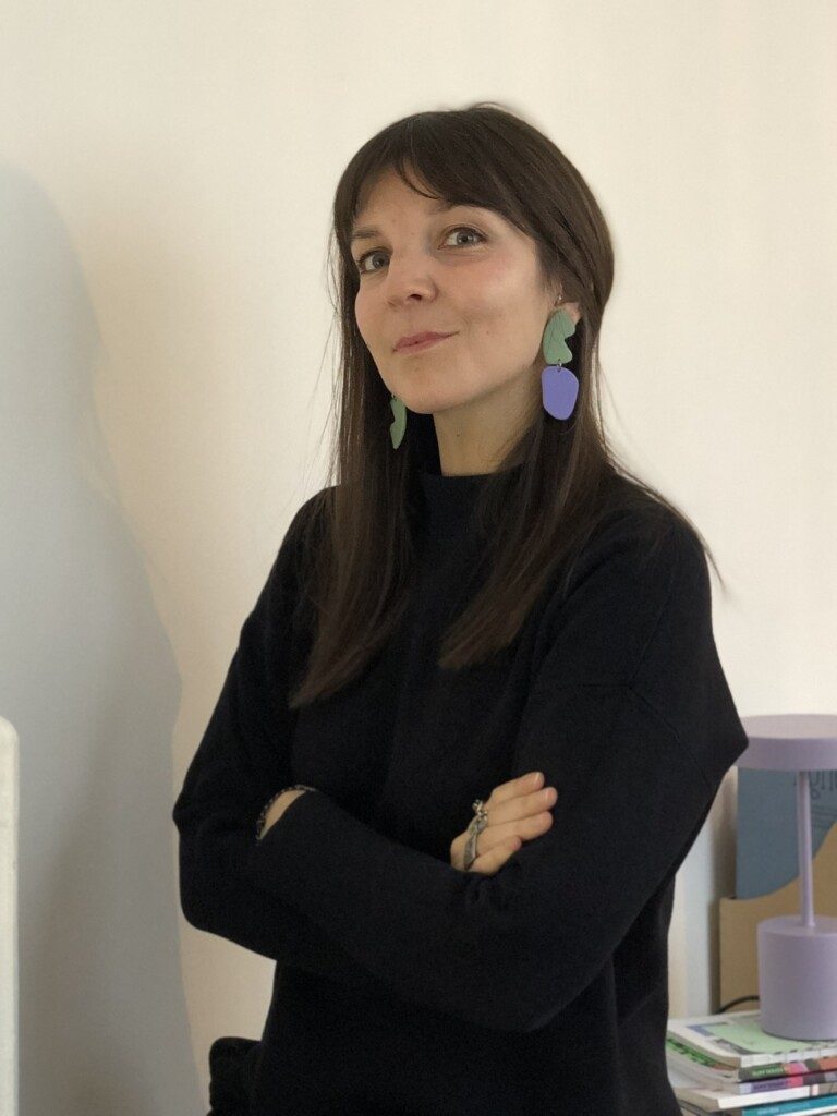 Portrait de l'autrice et chercheuse Emmanuelle Durand. Elle se tient debout, les bras croisés et porte un pull en laine noir et des boucles d'oreilles colorées vertes et violettes. 