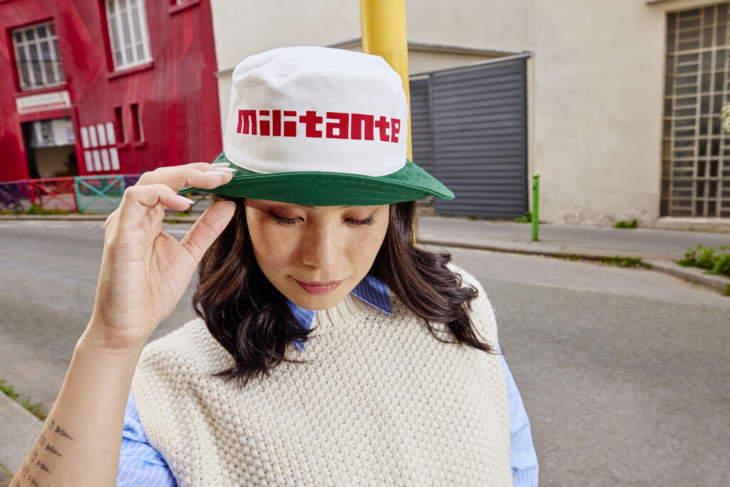 Une mannequin porte une casquette beige. Au centre, on peut lire en lettres rouges “militante"