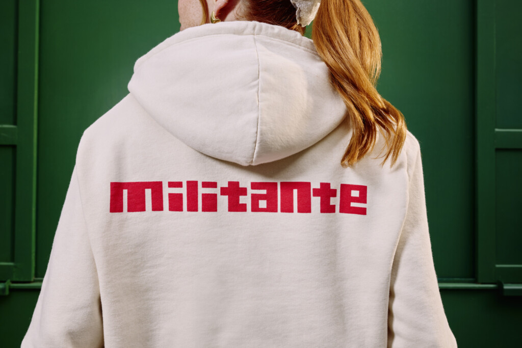 Une mannequin de dos porte un sweat à capuche beige. Sur le dos, on peut lire en rouge "militante"