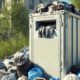 a container that usually in France receive used clothes. It's in the center of the image in a hyperrealistic type and 1920 x 700 px. In front, there is many bags piled up, themselves containing used clothes